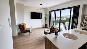 Een moderne woonruimte met een flatscreen-tv, een bank, een eethoek met een tafel, en een balkon met uitzicht over de stad.