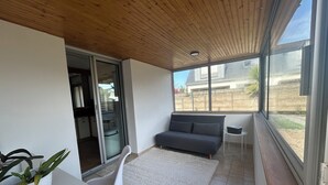 Ett modernt rum med en grå soffa, en vit stol, ett litet bord med en krukväxt och utsikt över ett hus genom stora fönster.