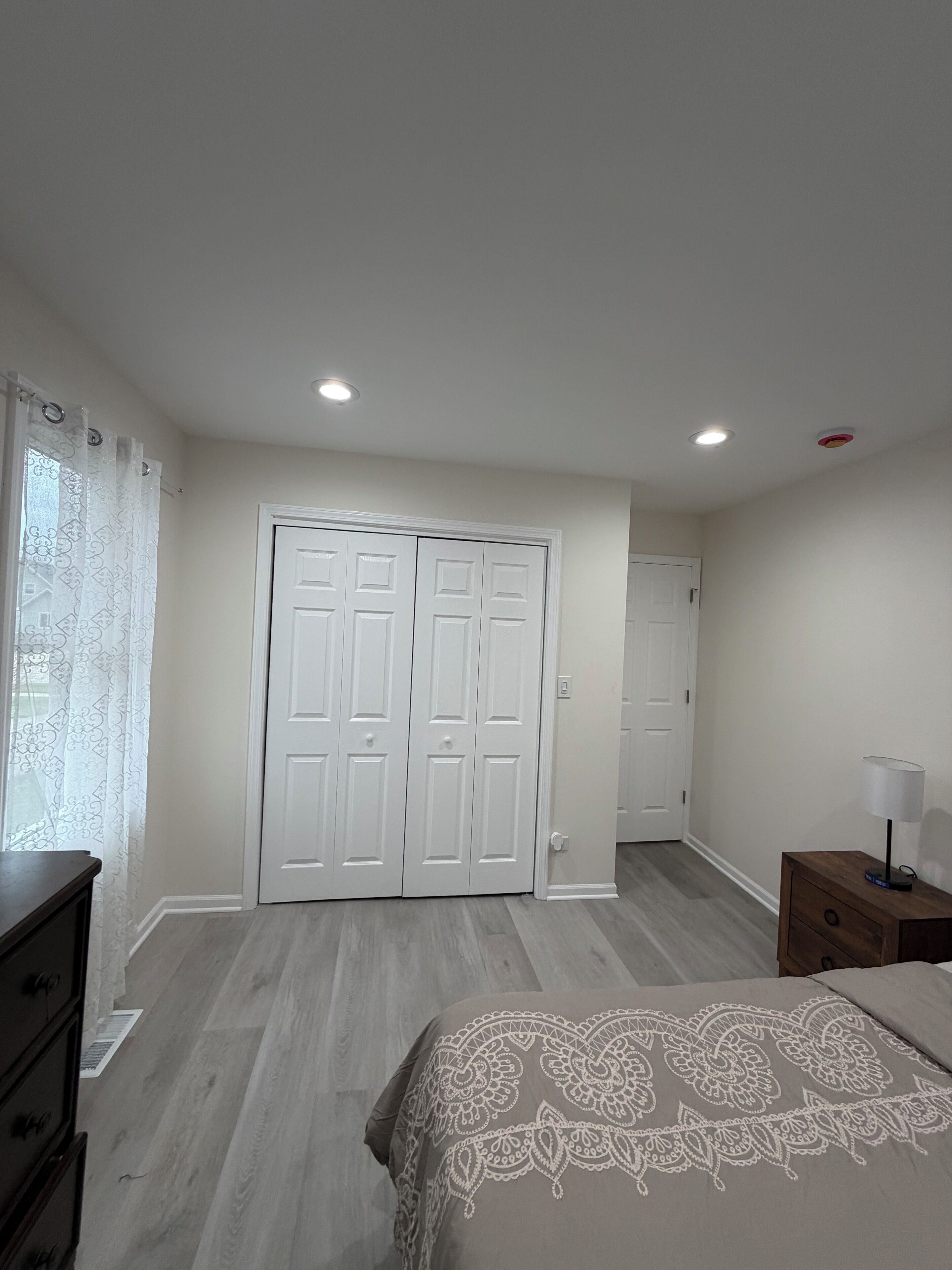 Une chambre à coucher comprenant un lit, une commode, une table de chevet, un placard à deux portes et une fenêtre avec un rideau.