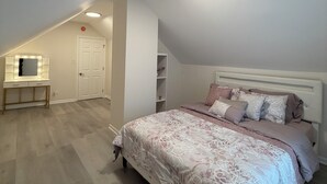 Un dormitorio con techo inclinado, una cama con ropa de cama estampada, un tocador con luces y una puerta blanca.