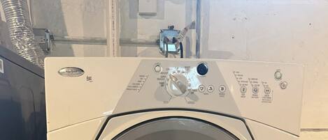 A front-loading washing machine with a control panel, located in a room with a concrete floor and a white wall.
