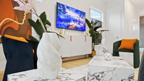 Ein modernes Wohnzimmer mit einer weißen Vase, einem Fernseher und einem Sofa mit einem orangefarbenen Kissen.