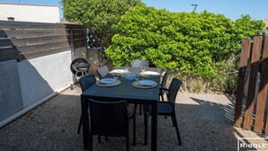 Un patio al aire libre con una mesa lista para comer, rodeada de sillas, una parrilla y un seto.