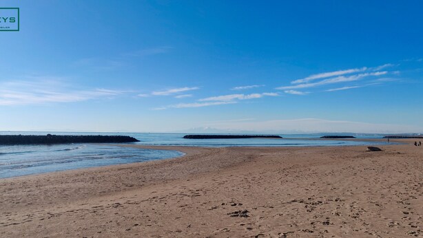 Una amplia playa de arena con aguas tranquilas, un rompeolas rocoso y montañas lejanas bajo un cielo despejado.