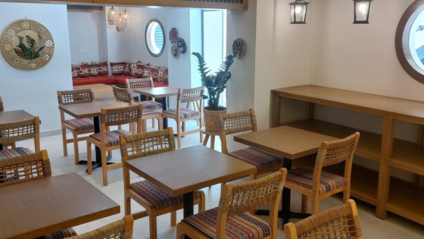 A dining area with wooden tables and chairs, a red sofa, decorative wall panels, and a large window. - B Suites Boutique Hotel (Amman)