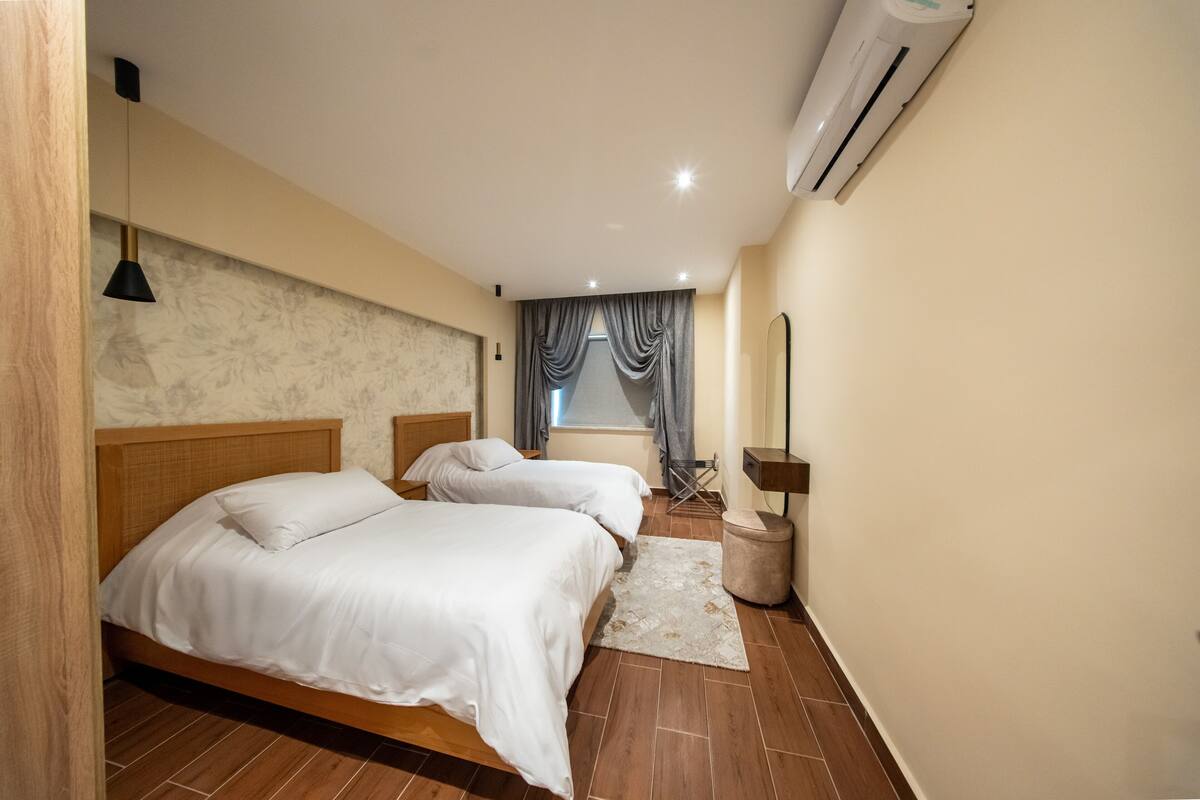 Une chambre avec deux lits, une tête de lit en bois, un petit tapis, un appareil de climatisation et une fenêtre avec des rideaux.