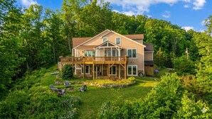 Une maison à deux étages dotée d’une terrasse, entourée d’une végétation luxuriante et d’arbres.