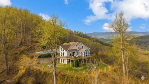 Une grande maison dotée d’une terrasse, entourée d’arbres et de collines.