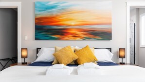 Un letto rifatto con cura, lenzuola bianche e azzurre, un dipinto al tramonto sulla parete e due abat-jour.