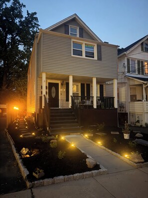 Een huis met twee verdiepingen, een veranda, wenteltrap naar de deur en een tuin met verlichting.