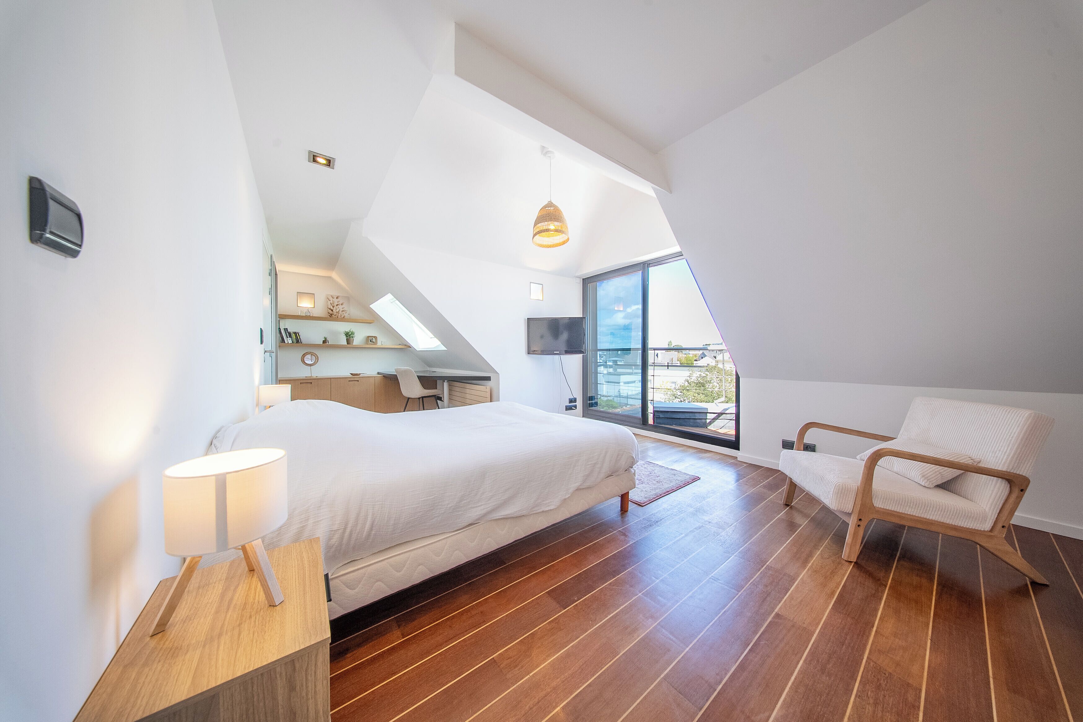 A modern bedroom with a large bed, a wooden nightstand, a chair, and a desk with a TV.