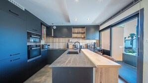A modern kitchen with dark cabinetry, a central island, and a large window. - Villa pieds dans l'eau - vue mer & jacuzzi (Larmor-Plage)