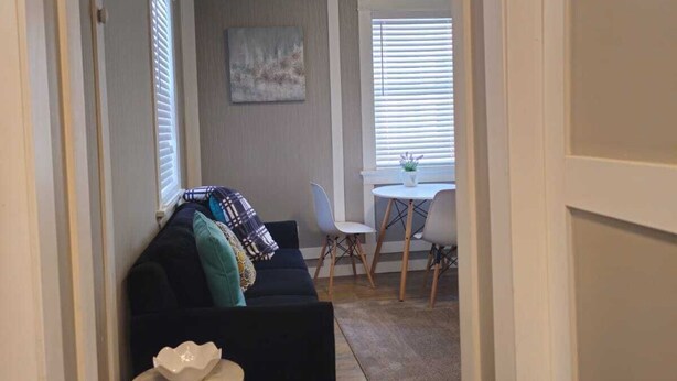Olohuoneessa on tummanvärinen sohva, pieni pyöreä pöytä, jonka päällä on valkoinen kulho, sekä ruokailutila, jossa on valkoinen pöytä ja tuolit.