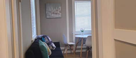 Ett vardagsrum med en soffa i mörk färg, ett litet runt bord med en vit skål och en matplats med ett vitt bord och stolar.