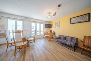 Ein Wohnzimmer mit Esstisch aus Glas, Korbstühlen, einem farbenfrohen Sofa, einem Fernseher und einem Deckenventilator.