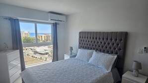 A bedroom with a large bed, a nightstand, a dresser, and a view of buildings through a window. - Stunning new Beachfront Condo 9th Floor Ocean View (Jacó)