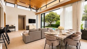 Una sala moderna con un sofá modular, una mesa de comedor preparada para seis comensales y un amplio ventanal con vistas a una piscina y follaje.