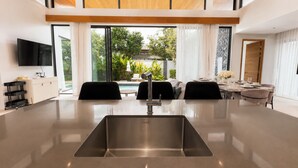 A modern kitchen with a central island, a dining area with a table set for four, and a large window offering a view of greenery. - Villa Luma Botanica | Private Pool Villa Near Bang Tao Beach (Tambon Choeng Thale)