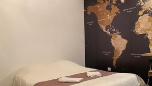 Una cama individual con una manta marrón y dos toallas dobladas, una pared oscura con un mapa mundial, y un piso de madera.