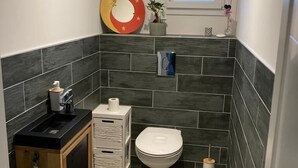 Un baño pequeño y funcional con inodoro, lavamanos y un armario de almacenamiento, con azulejos oscuros y un ventanal.
