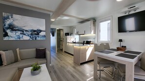 Un salon moderne comprenant un canapé, une table basse avec une plante, un coin repas avec une table et des chaises, une cuisine avec un îlot central et un téléviseur fixé au mur.