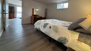 A bedroom with a bed, a desk, a chair, a dresser, a window, and a door leading to another room.