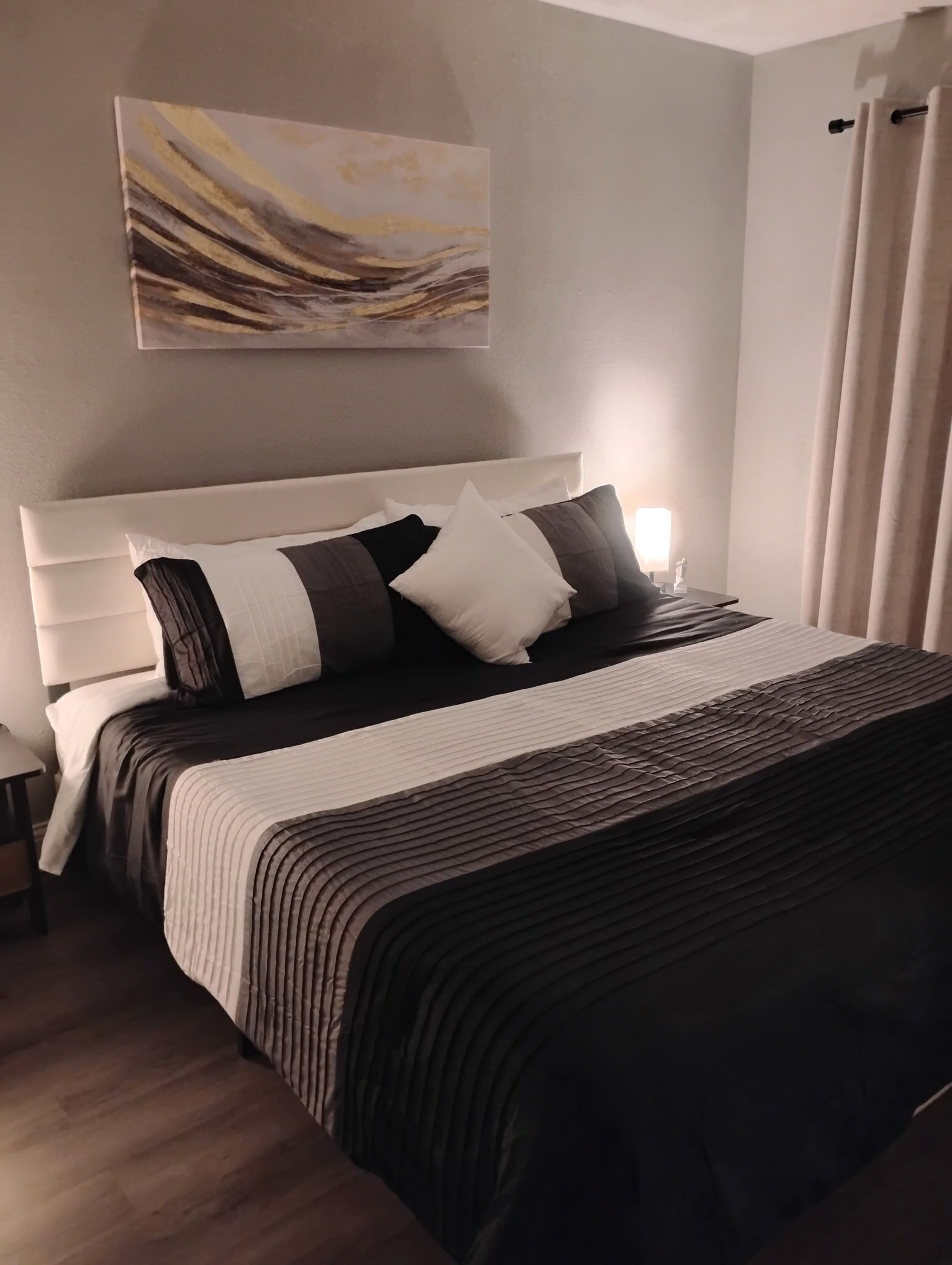 A neatly made bed with a striped bedspread and pillows, a white headboard, a bedside table with a lamp, and a painting on the wall.