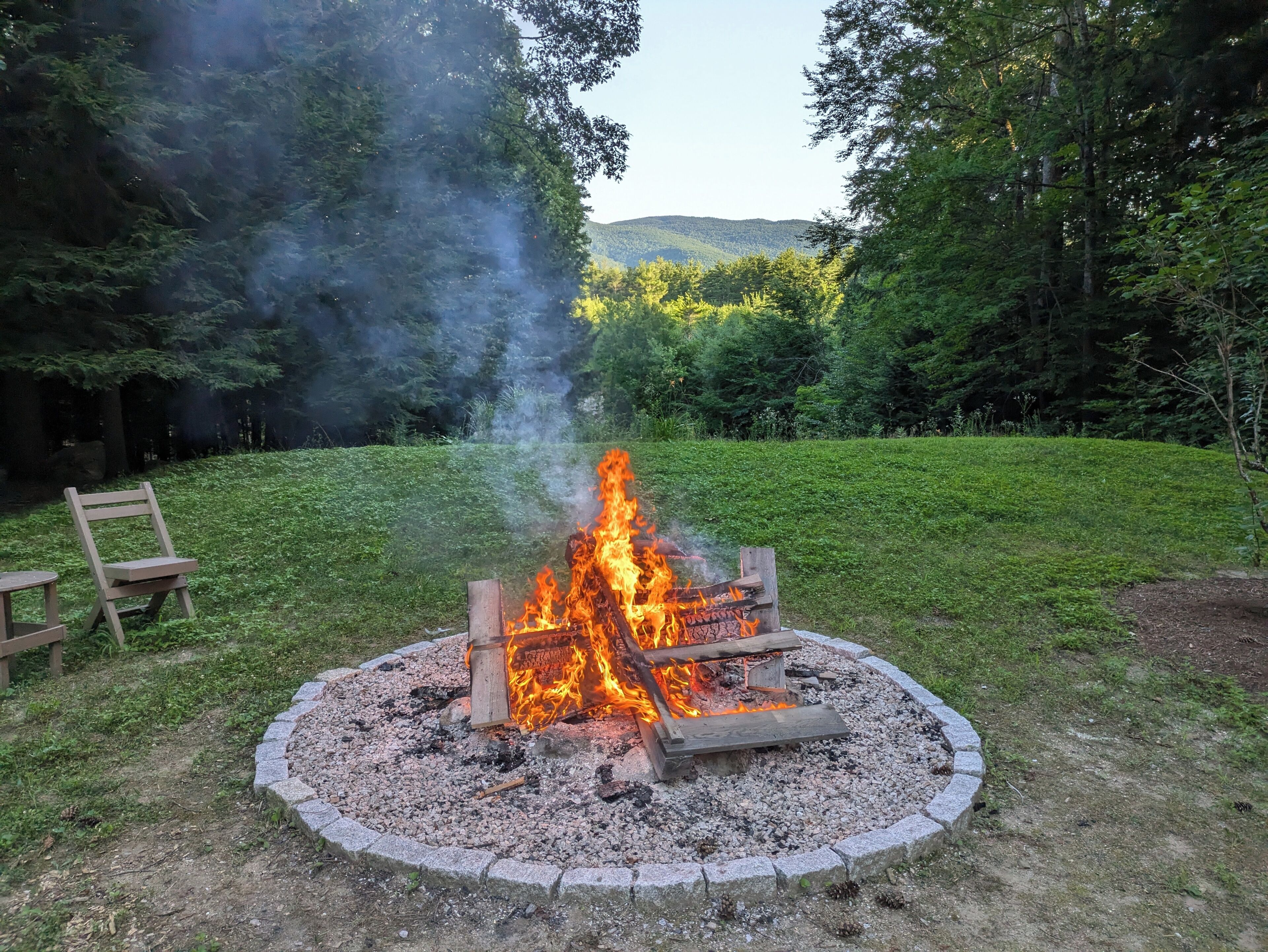 A circular fire pit with logs, surrounded by a stone border, in a grassy area with trees and hills in the background.
