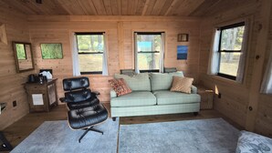 Interior de una cabaña de madera con un sofá, un sillón, una mesita y dos ventanas.