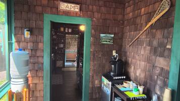 A rustic interior with a brick wall, a sign reading 'LAMP,' a water dispenser, a coffee machine, and a table with cups and utensils.