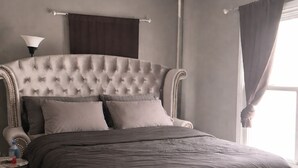 Een slaapkamer met een gestoffeerd hoofdeinde, een nachtkastje met een lamp, een waterfles en een raam met gordijnen.