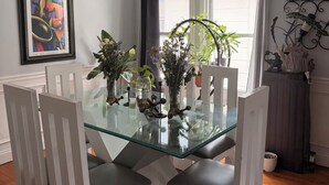 Een eetgedeelte met een glazen tafel, witte stoelen, een plafondventilator en potplanten.