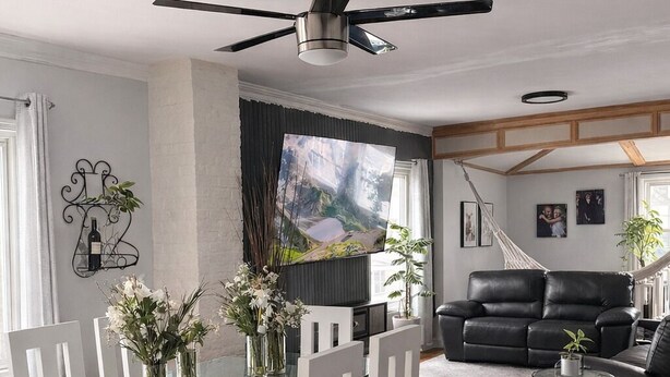 Een moderne woonkamer met een glazen eettafel, witte stoelen, een zwarte leren bank en een plafondventilator.