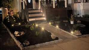 Een tweelaags huis met een voortuin, een veranda, trappen en een kleine tuin met verlichting.