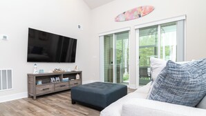 Una sala de estar con un televisor de pantalla plana, un reposapiés oscuro, un sofá de color claro y un ventanal que da al exterior.