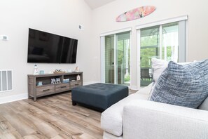 A living room with a flat-screen TV, a dark ottoman, a light-colored sofa, and a glass door leading outside. - Welcoming 4BR Villa with Pool in Charming Frankford (Frankford)