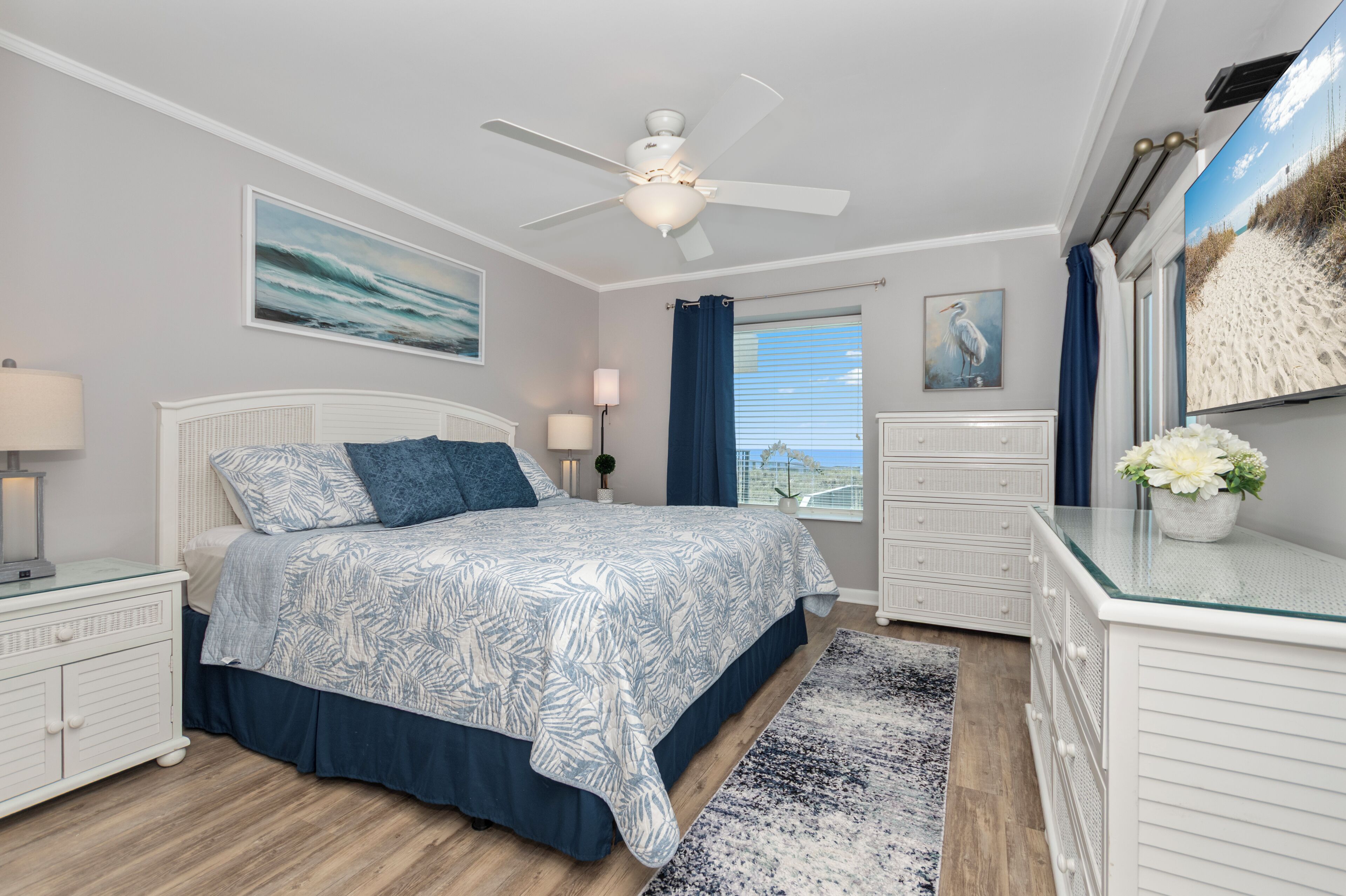 A bedroom with a bed, a ceiling fan, a window with curtains, a dresser, and a wall-mounted TV.