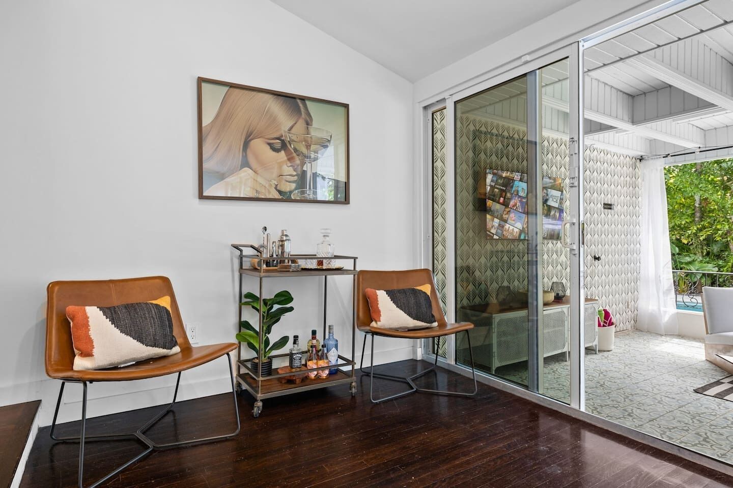 A modern room with two wooden chairs, a metal side table, a glass door leading to an outdoor area, and a large framed picture on the wall.