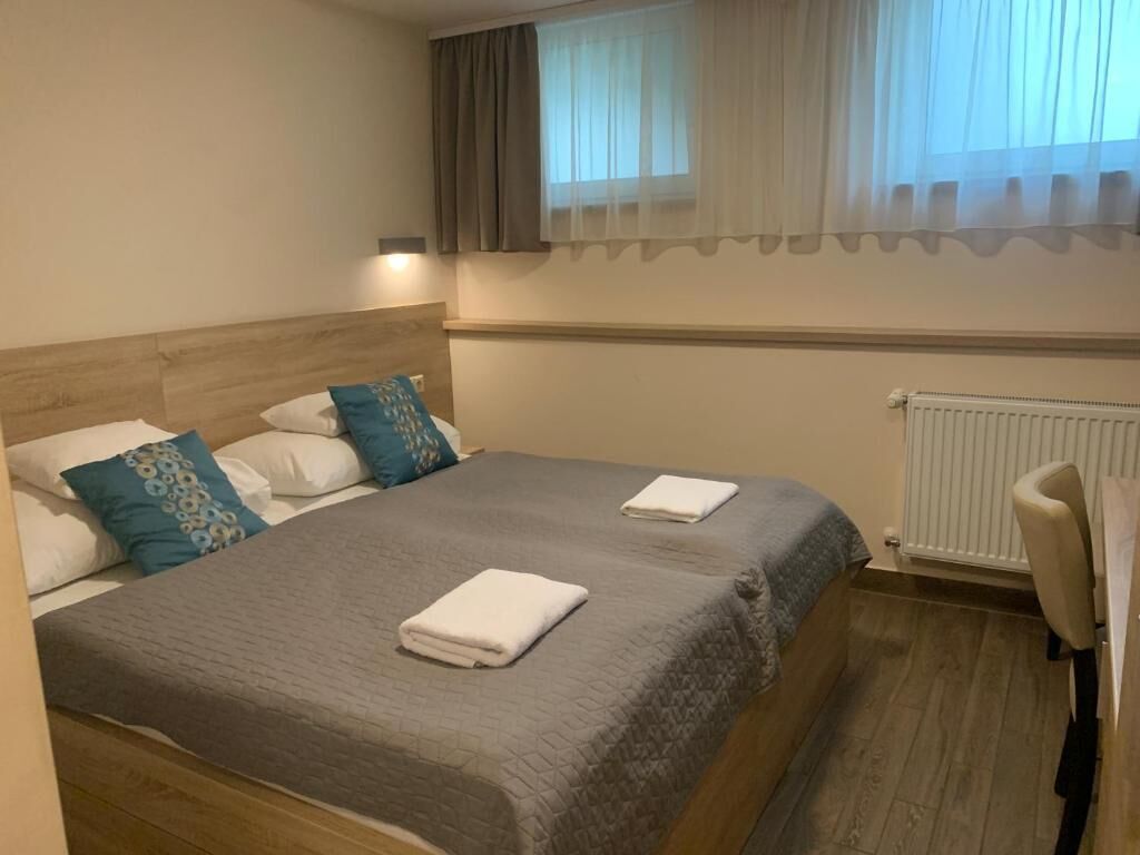 Ein ordentlich bezogenes Bett mit Kissen, ein Heizkörper und ein Sessel in einem Zimmer mit Fenster und Vorhängen.