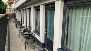 Un passadís d'hotel amb una fila de finestres, una porta i zona d'estar a l'exterior amb taules i cadires.