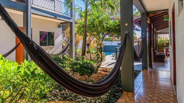 A hammock in a courtyard with a pool, a covered walkway, and a building with balconies. - Pousada papaya (Bonito)