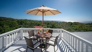 Ein runder Tisch mit vier Stühlen, ein beiger Sonnenschirm und farbenfrohe Getränke auf einem Balkon mit weißer Reling und schöner Aussicht.