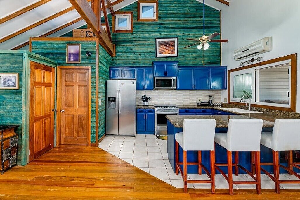 A kitchen with blue cabinets, a refrigerator, a microwave, and a bar area with white chairs.