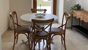 A dining area with a round table and four chairs, a view through a window, and a small shelf with a plant. - Il Villino - Luxury Country House, a luxury retreat between nature and tradition (Puglia)
