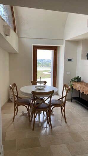 A dining area with a round table and four chairs, a view through a window, and a small shelf with a plant. - Il Villino - Luxury Country House, a luxury retreat between nature and tradition (Puglia)