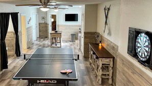 Ein Tischtennistisch mit Dartscheibe und Deckenventilator in einem Raum mit Holzboden und an der Wand montiertem Fernseher.