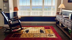 Ein Zimmer mit Erkerfenster, einem Schaukelstuhl, einem Schreibtisch mit Lampe und einem gemusterten Teppich.