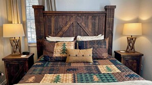 Ein Bett mit gesteppter Bettdecke, mehreren Kissen und einem Holzkopfende. Es gibt zwei Nachttische mit Lampen.
