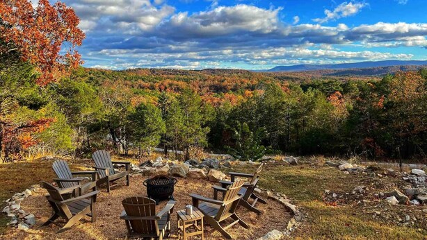 Ein runder Sitzbereich mit Adirondack-Stühlen und einer Feuerstelle, umgeben von einer natürlichen Landschaft mit Bäumen und Blick auf weit entfernte Hügel.