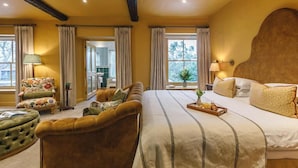A hotel room with a large bed, a sofa, a chair, and a small table. There are curtains on the windows and a lamp on the side table.